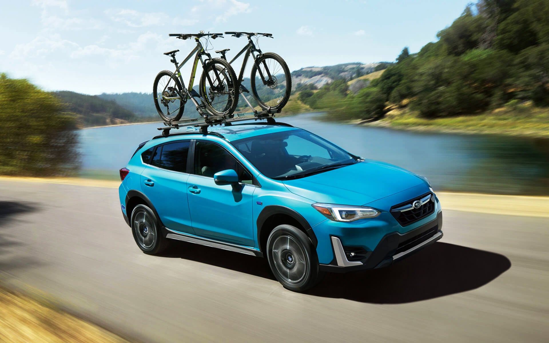 A blue Crosstrek Hybrid with two bicycles on its roof rack driving beside a river | Royal Moore Subaru in Hillsboro OR
