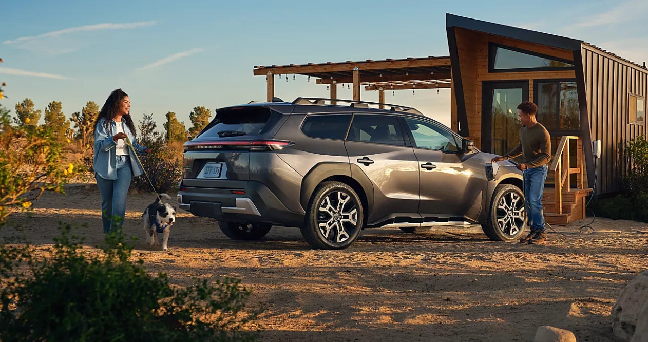 A gray silver Subaru Trailseeker parked on sand by a small shack with a woman walking her dog behind it and a man standing in front of it.