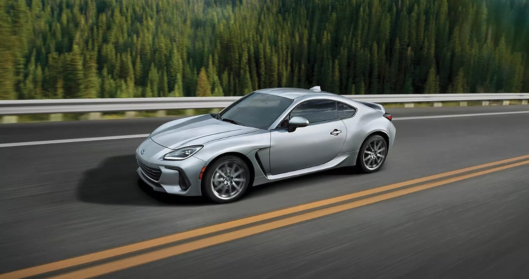 A silver Subaru BRZ driving down the road by a forest.
