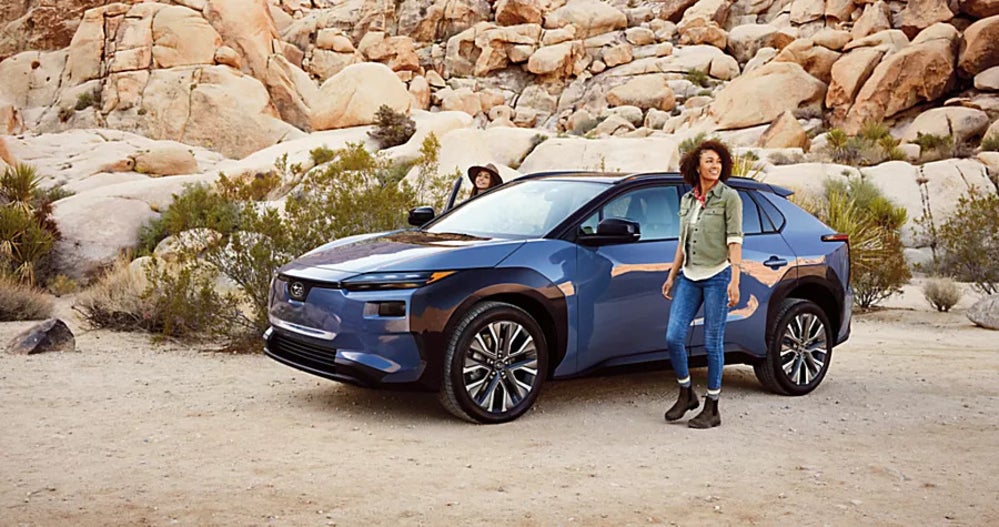 A blue Subaru Solterra parked by a rocky outcrop off-road, while a pair of women get out of it.